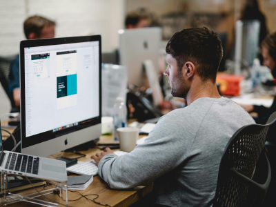 Ein Manager in einem grauen Pullover sitzt vor einem iMac und arbeitet