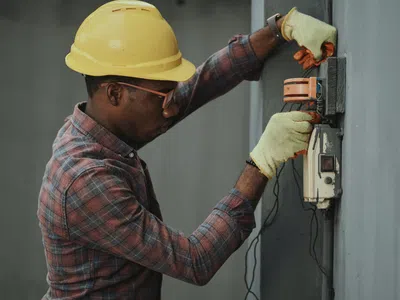 Techniker:in Elektrotechnik mit Schutzhelm und Schutzhandschuhen arbeitet an einer Wand.