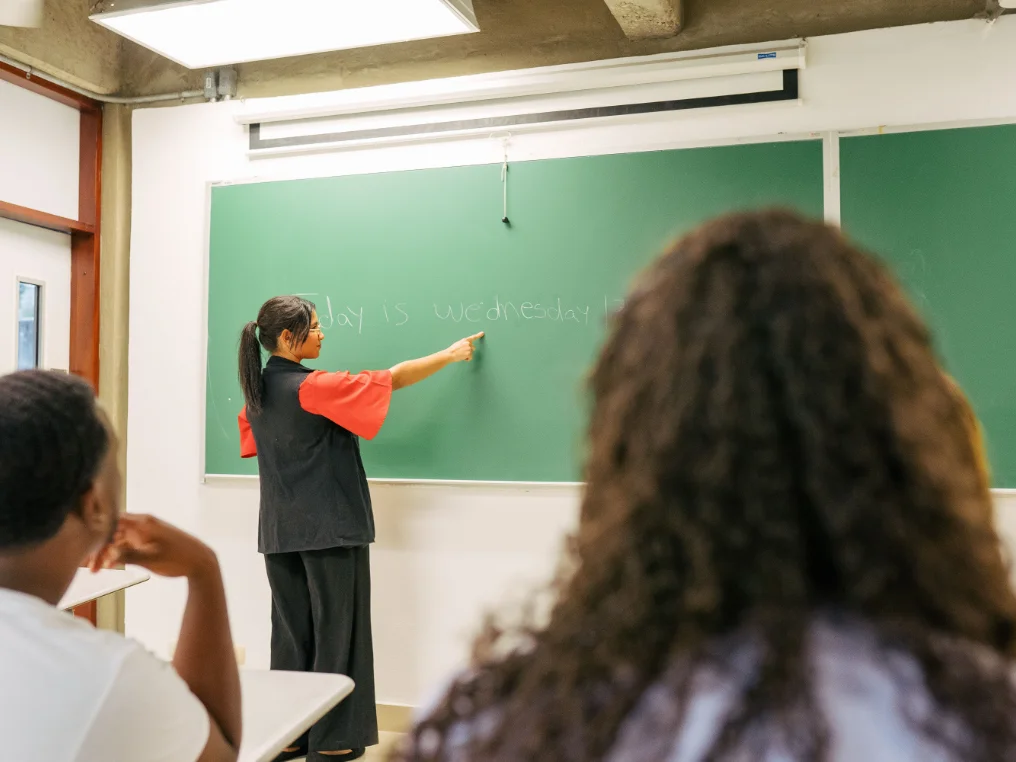 Eine Englischlehrerin steht an der Tafel und zeigt einen Satz zwei Schülern