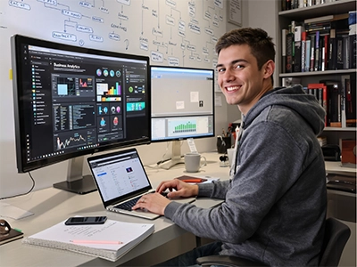 Ein Wirtschaftsinformatik-Student sitzt am Schreibtisch vor einem Laptop und zwei großen Bildschirmen.