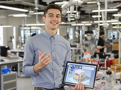 Ein Projektmanagement-Student steht in einem großen Kreativraum und stellt sein Projekt mithilfe eines Tablets vor.  
